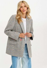 Hnědý houndstooth blazer s multibarevnými fleky. Má růžové knoflíky, oversized klopy a volný střih. Nositelný přes bílý top a modré džíny.