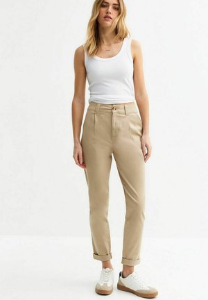 Pantalons beiges taille haute avec une jambe fuselée, ourlets à revers et poches avant, associés à un débardeur blanc et des baskets blanches.