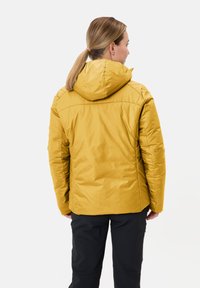 Giacca gialla imbottita con cappuccio, caratterizzata da una texture liscia e dettagli delle cuciture. Indossata sopra pantaloni neri, mostrando la vista posteriore.