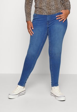 Kvinde iført blå skinny jeans, hvide højtop-sneakers og en langærmet top med leopardprint, stående med den ene hånd på hoften.