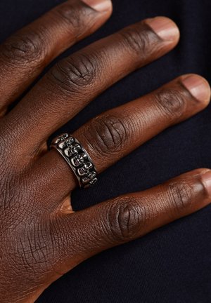 Zwarte metalen ring met een gestructureerd oppervlak, gekenmerkt door een rij ronde accenten. Draagt op een hand met een diepe huidskleur, waarbij de details van de vingers zichtbaar zijn.