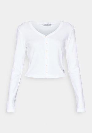 Weißer, gekürzter Cardigan mit V-Ausschnitt, langen Ärmeln und Knopfverschluss vorne. Hat eine weiche Textur und ein kleines Logoshirt am Saum.