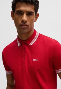 Rotes Polohemd aus strukturiertem Stoff, mit einem gestreiften Kragen und einem weißen, gestickten "BOSS"-Logo auf der linken Brust.