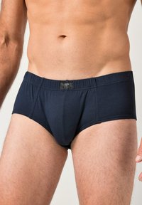 Torace maschile e parte superiore delle cosce indossando slip aderenti blu scuro con un piccolo logo frontale sulla fascia elastica in vita.