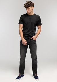 Młody mężczyzna z kręconymi włosami, ubrany w czarną koszulkę, wyblakłe czarne jeansy i niebieskie trampki, stoi na tle jednolitego jasnego tła.