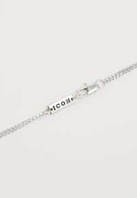 Bracelet en chaîne argentée avec une étiquette rectangulaire gravée du mot « ICON » et un fermoir en mousqueton. Texture lisse et finition polie.
