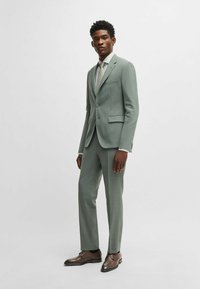 Completo su misura verde chiaro con giacca slim fit dotata di rever a punta, chiusura con bottone singolo e pantaloni con tasche piatte. Scarpe eleganti marroni.