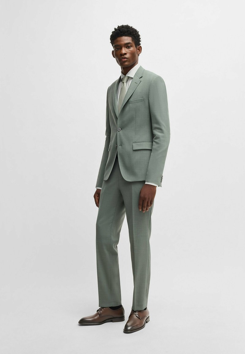 Completo su misura verde chiaro con giacca slim fit dotata di rever a punta, chiusura con bottone singolo e pantaloni con tasche piatte. Scarpe eleganti marroni.