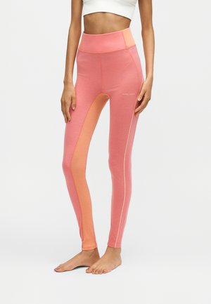 Mujer con leggings bicolor de talle alto en rosa y coral y un top corto blanco, de pie descalza sobre un fondo neutro.