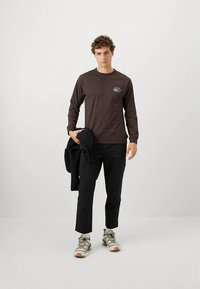 Braunes Langarmshirt mit kleinem Motiv auf der Brust, kombiniert mit schwarzen Hosen und grauen Turnschuhen. Das Modell hält eine schwarze Fleecejacke in der Hand.