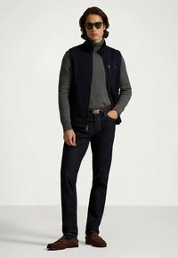 Vīrietis ar saulessargiem, tumšā bezdiega veste, pelēks turtlenecks svīteris, tumši zili džinsi un brūni loaferi stāv ar vienu roku kabatē.