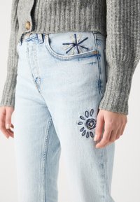 Lyseblå denimjeans har øyebroderi og et stjerneskuddsdesign nær framlommen, parret med en grå strikkegenser.