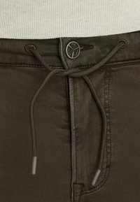 Un pantalon vert olive foncé avec un cordon de serrage et un bouton en métal avec un logo embossé, accompagné de poches avant plates.