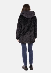 Cappotto in ecopelliccia nera con un cappuccio liscio e trapuntato. Indossato sopra jeans di denim e stivali neri chunky. Texture morbida con una vestibilità morbida.
