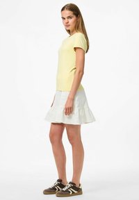 Geel T-shirt met korte mouwen, gecombineerd met een witte rok met ruches en donkerbruine sneakers met lichte accenten, getoond op een model.