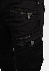 Pantaloni cargo neri con diversi design di tasche, accessori metallici e tessuto testurizzato. Cerniere e dettagli del logo ben visibili.