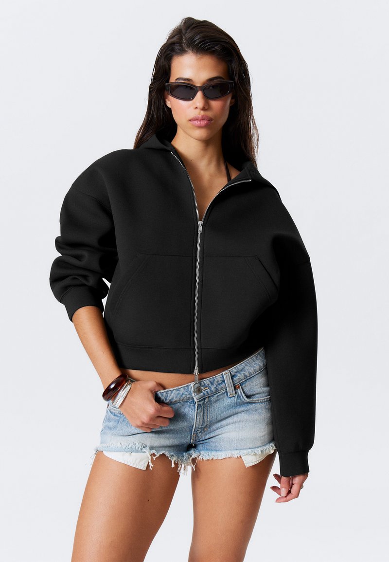 Sweatshirt noir court avec fermeture éclair devant et grande poche, associé à un short en denim clair déchiré. Tissu lisse et épais avec une coupe décontractée.