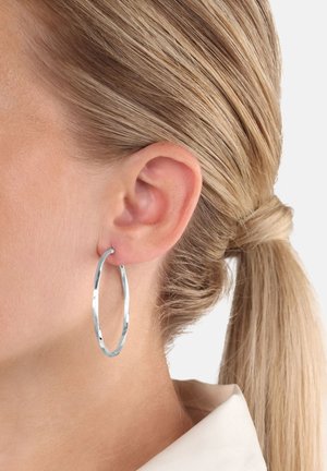Primer plano de la oreja de una mujer con un pendiente en forma de aro retorcido de plata y el cabello rubio recogido en una coleta.
