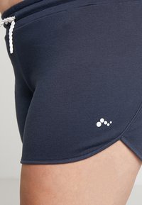 Pantalones cortos deportivos de color azul marino fabricados con tejido elástico, con una cintura ajustable mediante cordón y un pequeño logo blanco ubicado en el lado izquierdo.