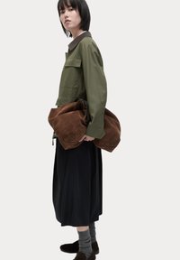 Olijfgroene jas met een bruine kraag, holding een grote bruine suède tas, zwarte rok, en grijze sokken, terwijl ze donkere schoenen draagt.