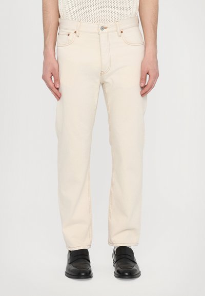 Jean en coton beige avec une coupe droite, cinq poches et des coutures contrastées, associés à des chaussures noires à enfiler.