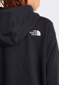Personne portant un sweat à capuche noir avec le logo The North Face brodé en blanc sur le haut du dos près de la capuche.