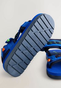 Blå sandaler med en tekstureret gummisål, justerbare stropper og farverige accenter. Har et zigzag grip-mønster på sålen.