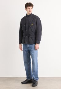 Camisola de algodão preta com colarinho alto, múltiplos bolsos frontais, cintura cingida, e um patch de logo, usada com jeans azul claro e sapatos pretos.