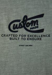 Camiseta de algodón verde grisáceo con texto impreso en negro: "Hecho a medida", "Elaborado para la excelencia, construido para perdurar" y "Street One Men".
