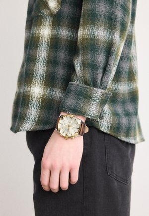 Grønn og grå rutete skjorte med rullede ermer, kombinert med et gull- og kremfarget klokke med brunt lærrem og kronografdetaljer.