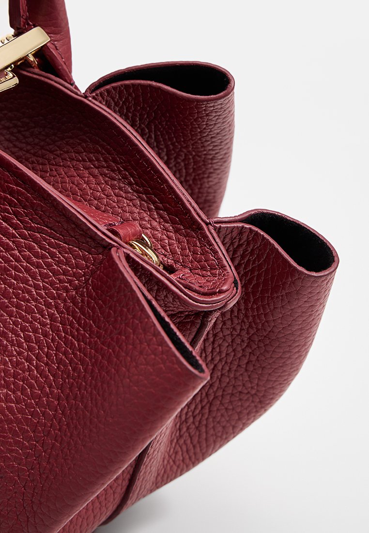 Borsa in pelle bordeaux con finitura testurizzata, caratterizzata da una chiusura dorata e un'apertura superiore. La forma è leggermente arrotondata con bordi svasati.