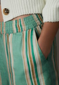 Pantalon à rayures vertes avec une ceinture texturée et une poche latérale, présentant des rayures verticales dans des nuances de vert, rose et beige.