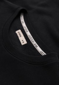 Gros plan sur un sweat-shirt en coton noir à col ras du cou, montrant le col côtelé et l'étiquette intérieure avec le nom de la marque et la taille moyenne.