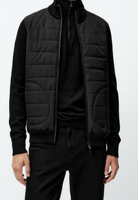 Homme portant un gilet matelassé noir par-dessus un haut zippé à manches longues noir et un pantalon noir, sur un fond uni.