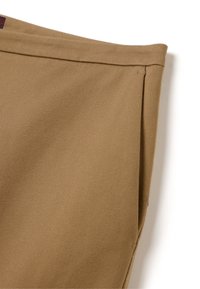 Gros plan sur un détail de poche en tissu beige sur un pantalon avec des coutures visibles et un bord plié sur fond blanc.