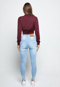 Bordowy, długi rękaw, krótki top zestawiony z jasnoniebieskimi, obcisłymi dżinsami z przetarciami na kolanach. Białe sneakersy i widoczna brązowa skórzana metka.