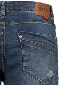 Donkerblauwe spijkerbroek van denim met een leren label met de tekst "SUBLEVEL", zilverkleurige hardware en zichtbare stiksels op de achterzakken.
