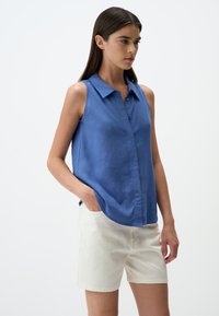 Helles blaues, ärmelloses Hemd mit Kragen und Knopfleiste. Getragen mit beigen Shorts. Glatte Textur, lässige Passform und ohne sichtbare Muster.