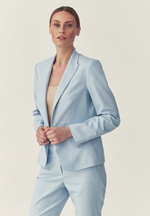 Kvinde med håret bundet tilbage iført en lyseblå jakkesætjakke og bukser over en beige top, stående mod en ensfarvet baggrund.