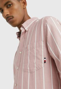 Camicia button-up rosa chiaro con strisce verticali bianche, taschino sul petto e toppa con logo nei colori rosso, bianco e blu.