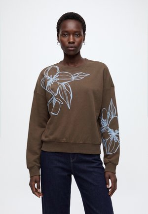 Kvinde iført brun sweatshirt med store hvide blomsterlinjetegninger på brystet og venstre ærme, kombineret med mørkeblå jeans, står foran en hvid baggrund.