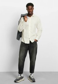 Light-colored button-up shirt worn with dark jeans. Model holds a gray jacket over one shoulder, wearing gray sneakers. Clean, casual styling.