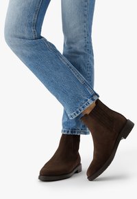 Persona vestida con jeans azules y botas Chelsea de ante marrón oscuro con tacones bajos, de pie con una pierna cruzada sobre la otra.