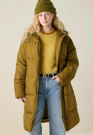Woman wearing olive green beanie and long quilted olive green coat over mustard sweater and light blue jeans, standing with one hand in pocket.