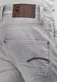 Vaqueros de mezclilla gris claro con parche de cuero marrón en la cintura, que cuentan con dos bolsillos traseros con detalles de costura y remaches de metal.