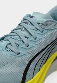 Αθλητικό παπούτσι σε ανοιχτό μπλε χρώμα με υφή στο επάνω μέρος, λεπτομέρειες από δίχτυ και μαύρες πινελιές. Διαθέτει επίπεδα κορδόνια και φωτεινή κίτρινη σόλα.