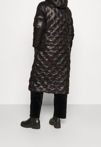 Abrigo negro largo y acolchado con capucha, con textura de edredón y acabado brillante. Combinado con pantalones de pana negros y botas gruesas negras.