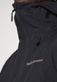 Casaco impermeável preto com colarinho alto, fecho na frente e cordões de ajuste. Apresenta o logótipo "PeakPerformance" em bordado branco no peito.