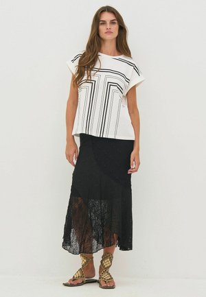 Femme aux longs cheveux portant un haut blanc à motif géométrique, une jupe en dentelle noire asymétrique et des sandales ornées de clous dorés, posant devant un fond uni.