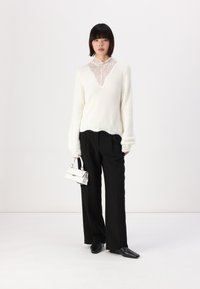 Pull blanc moelleux avec des détails en dentelle au col, associé à un pantalon noir à jambe large et à un petit sac à main blanc. Modèle portant des chaussures noires.